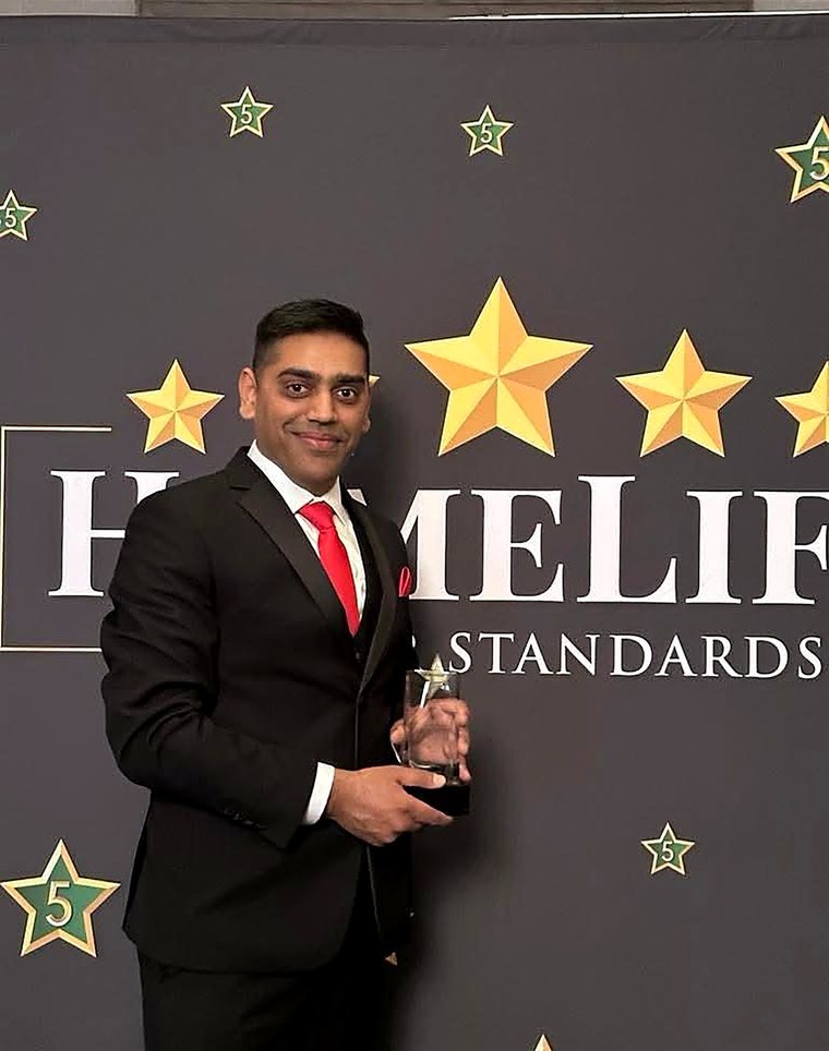 Robin Patel in a black suit holding a glass award in front of a HomeLife Higher Standards backdrop.